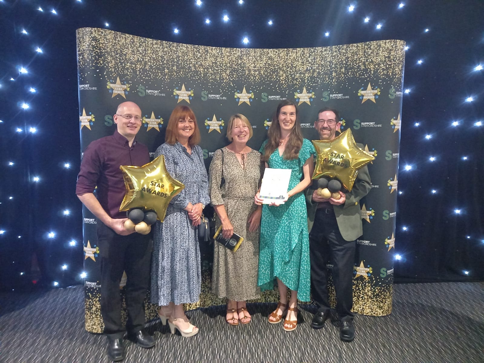 A group of people in front of a starry display board. They are holding a certificate and balloons