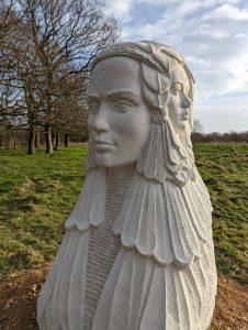 Stone white sculpture with goddess's face in foreground of picture, with grass, trees and sky shown in background.